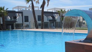 Una piscina con un moderno elemento acuático, rodeada de palmeras y edificios contemporáneos.