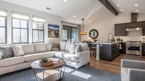 A spacious living room with a large L-shaped sectional sofa, a round coffee table, and a dining area with a kitchen in the background.