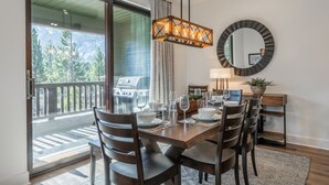 A dining area with a wooden table set for four, a chandelier, a round mirror, and a view of a balcony with a grill.