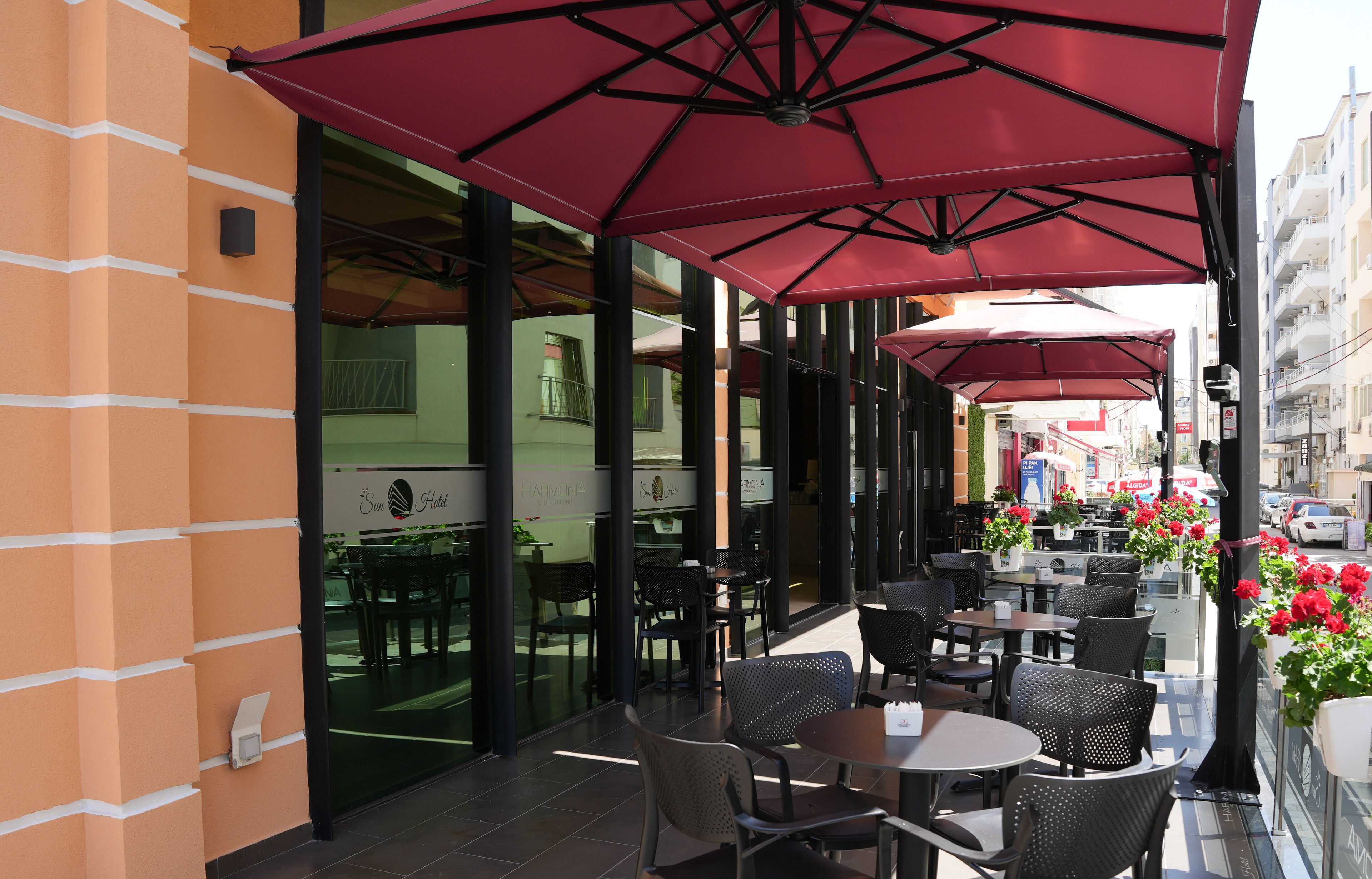 Un café en plein air avec des tables et des chaises sous des parasols rouges, un mur de verre et une vue sur la rue.