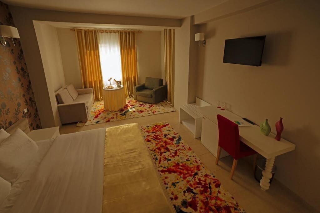 Ein Hotelzimmer mit Bett, Schreibtisch, Stuhl, Fernseher und Blick auf einen Balkon mit Vorhängen.