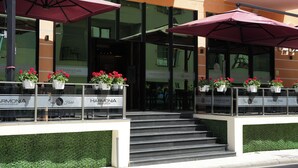 Fachada de un hotel con escalones que llevan a una puerta de cristal, zona de estar al aire libre con sombrillas y macetas con flores en la barandilla.