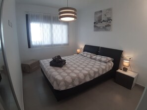 Un dormitorio con cama, dos mesitas de noche, una ventana con cortinas, una lámpara colgante y una obra de arte en la pared.