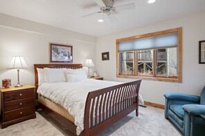 Un dormitorio con cama de madera, dos mesitas de noche, un velador, un sillón azul, una ventana con persianas, un ventilador de techo y un cuadro enmarcado en la pared.