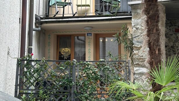 A two-story building with a balcony, a small table, and chairs. There is a potted plant on the balcony and a stone wall in the background. - Casa Edera  (Locarno)