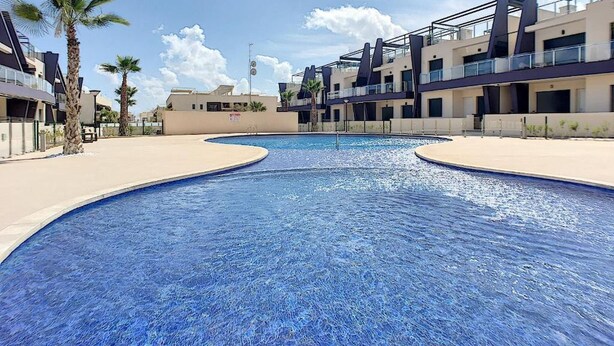 A swimming pool with a curved edge, surrounded by modern buildings with balconies and palm trees. - Penthouse Elisa Mil Palmeras (Torre de la Horadada)