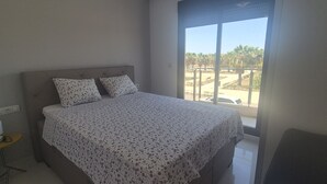 A bedroom with a bed, a nightstand, a lamp, and a window with a view of palm trees. - Penthouse Elisa Mil Palmeras (Torre de la Horadada)