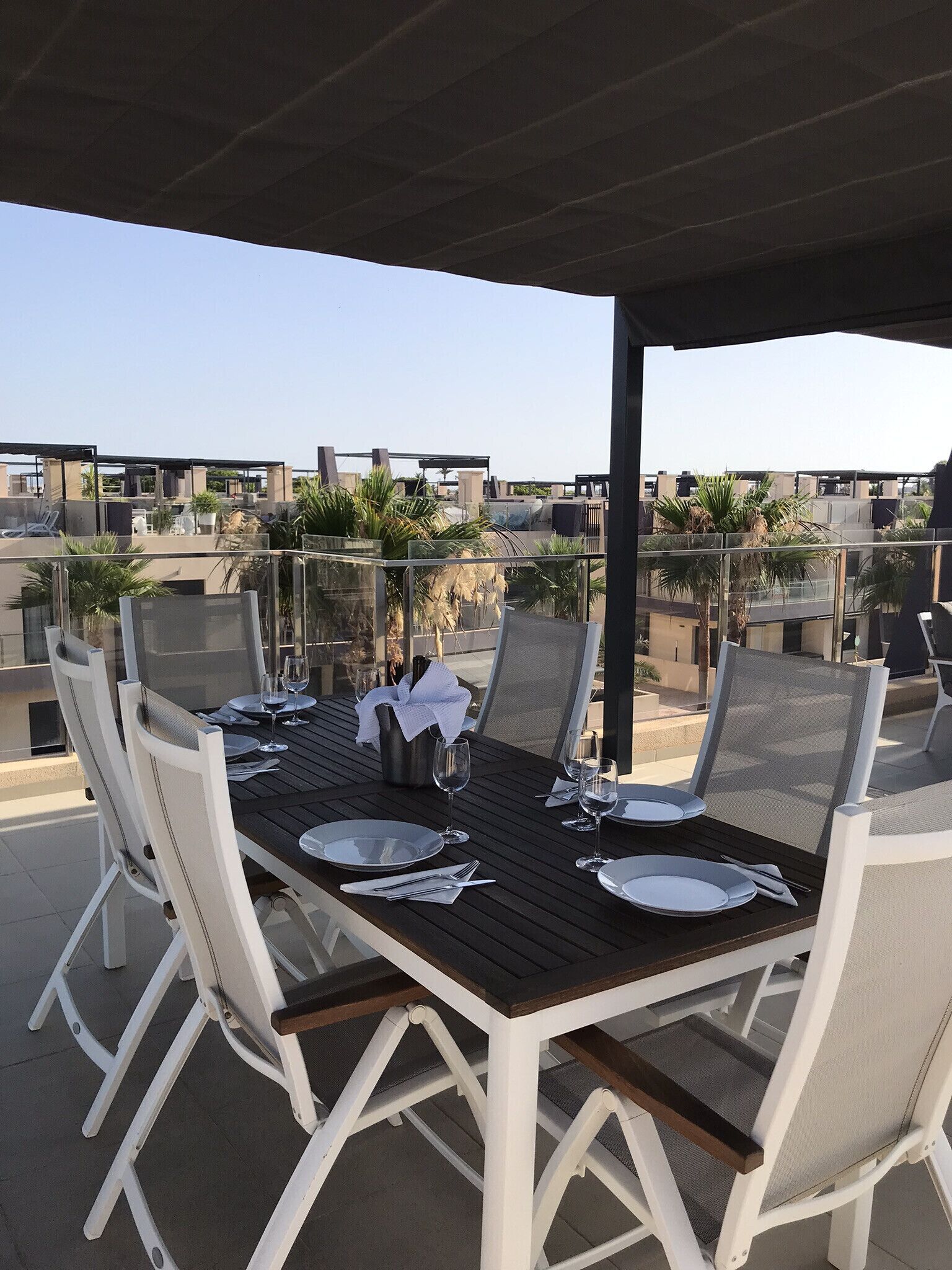 An outdoor dining area with a wooden table and white chairs, surrounded by glass railings and palm trees.