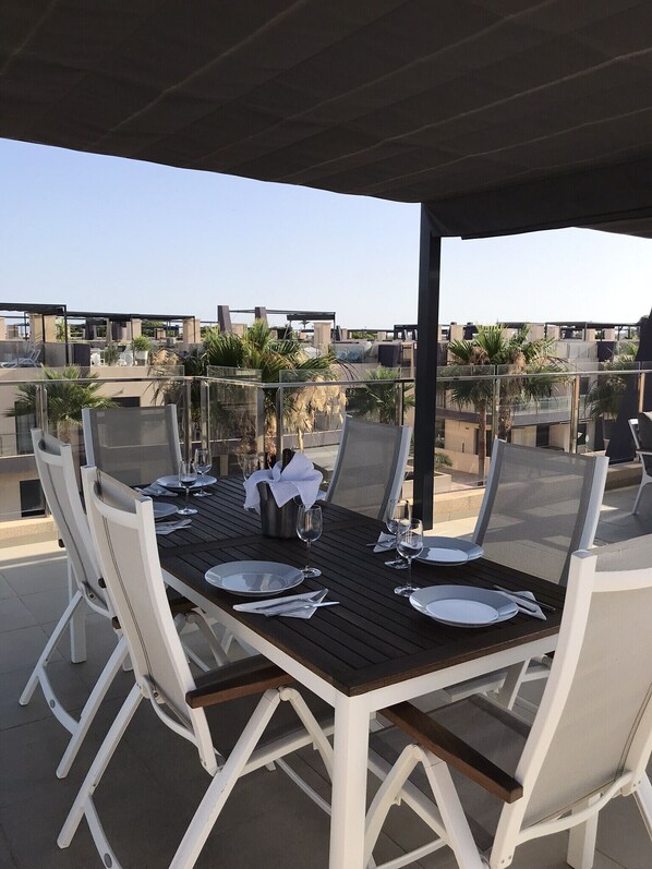 An outdoor dining area with a wooden table and white chairs, surrounded by glass railings and palm trees.