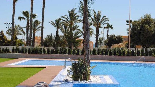 A swimming pool with a tiled deck, surrounded by palm trees and a white fence. - Apartment Aqua nature La mata; costa Blanca (La Mata)