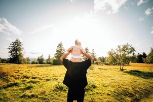 Una persona lleva a un niño en un campo cubierto de hierba, con árboles y cielo despejado.