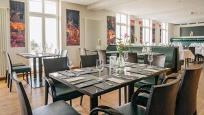 A restaurant with multiple dining tables and chairs, large windows, and abstract paintings on the walls.