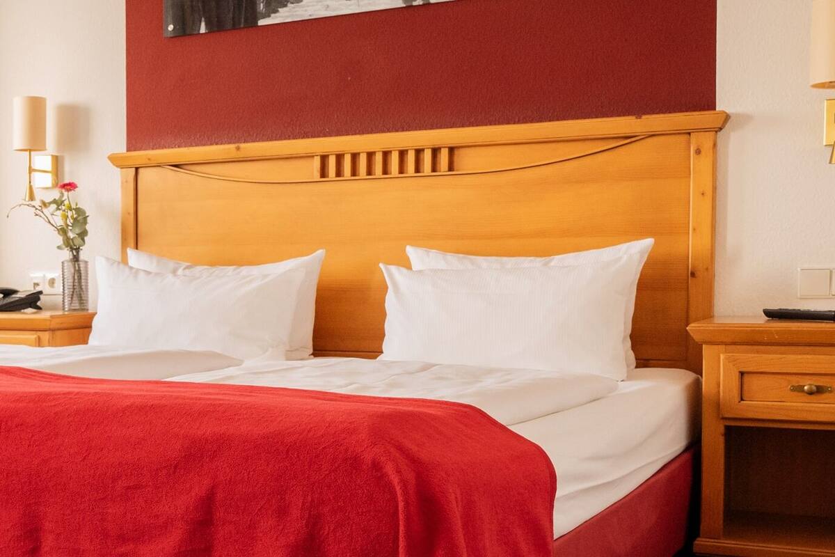 Une chambre d’hôtel avec un grand lit, une tête de lit en bois, une table de chevet avec une lampe et une fenêtre avec des rideaux.