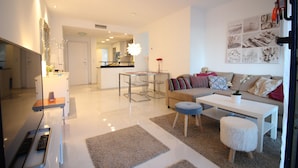 A modern living room with a sofa, coffee table, and ottoman. There is a dining area with a glass table and chairs. A kitchen is visible in the background. - Groundfloor Sea Senses Punta Prima , Orihuela Costa (Torrevieja)