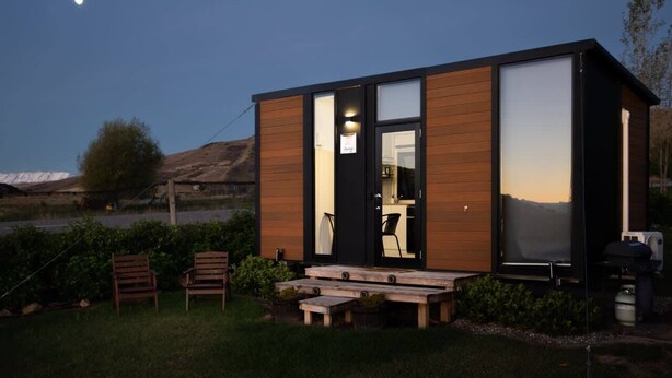 A modern cabin with large windows, a wooden exterior, and a patio area with chairs and a table. - Ruffell Ranch Cabin by Tiny Away (Tarras)