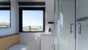 A bathroom with a toilet, a small cabinet, a window with a view, and a glass shower enclosure. - Ruffell Ranch Cabin by Tiny Away (Tarras)