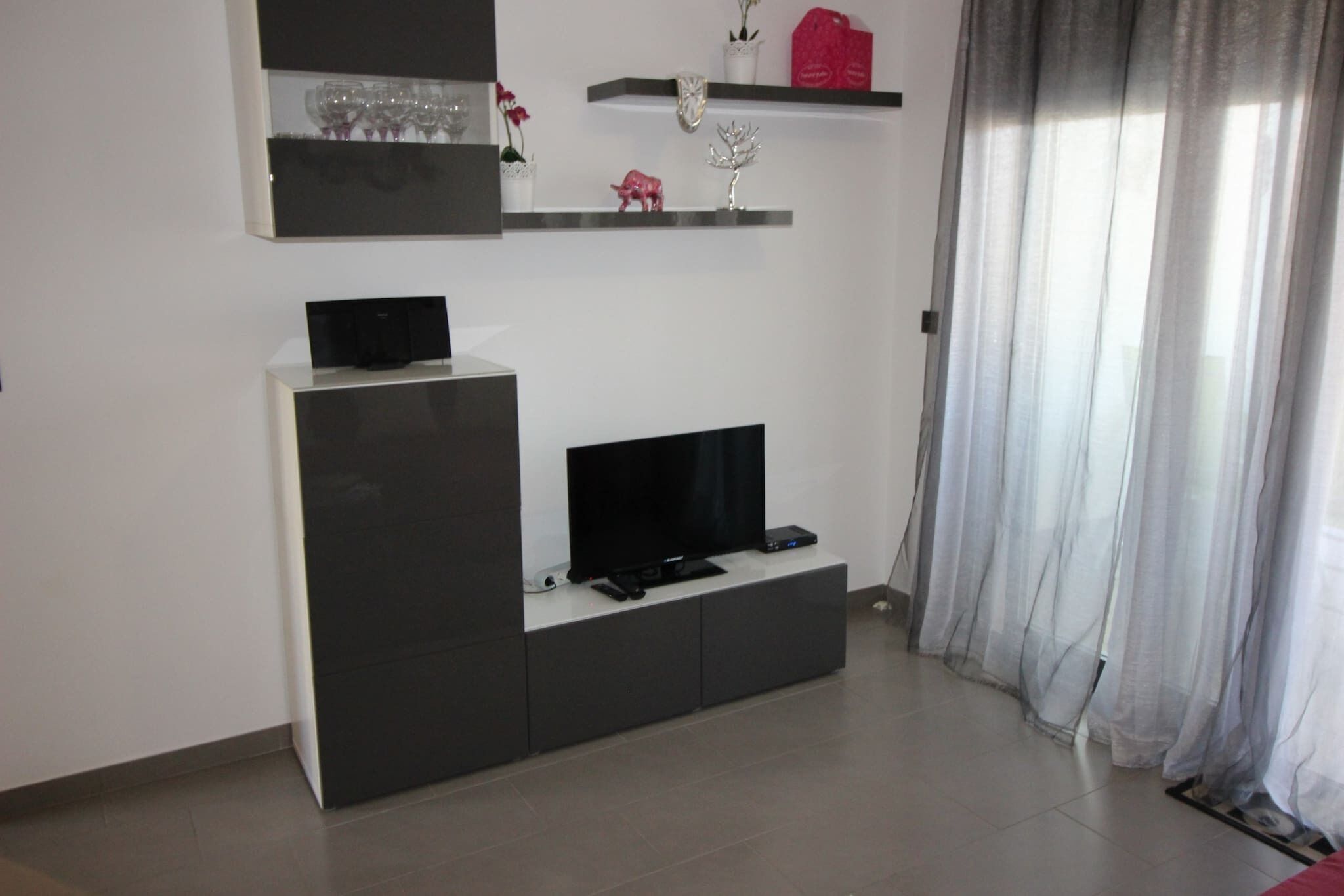 Een moderne woonkamer met een flatscreen televisie op een zwarte dressoir, een wit boekenrek met decoratieve objecten, en een raam met dunne gordijnen.