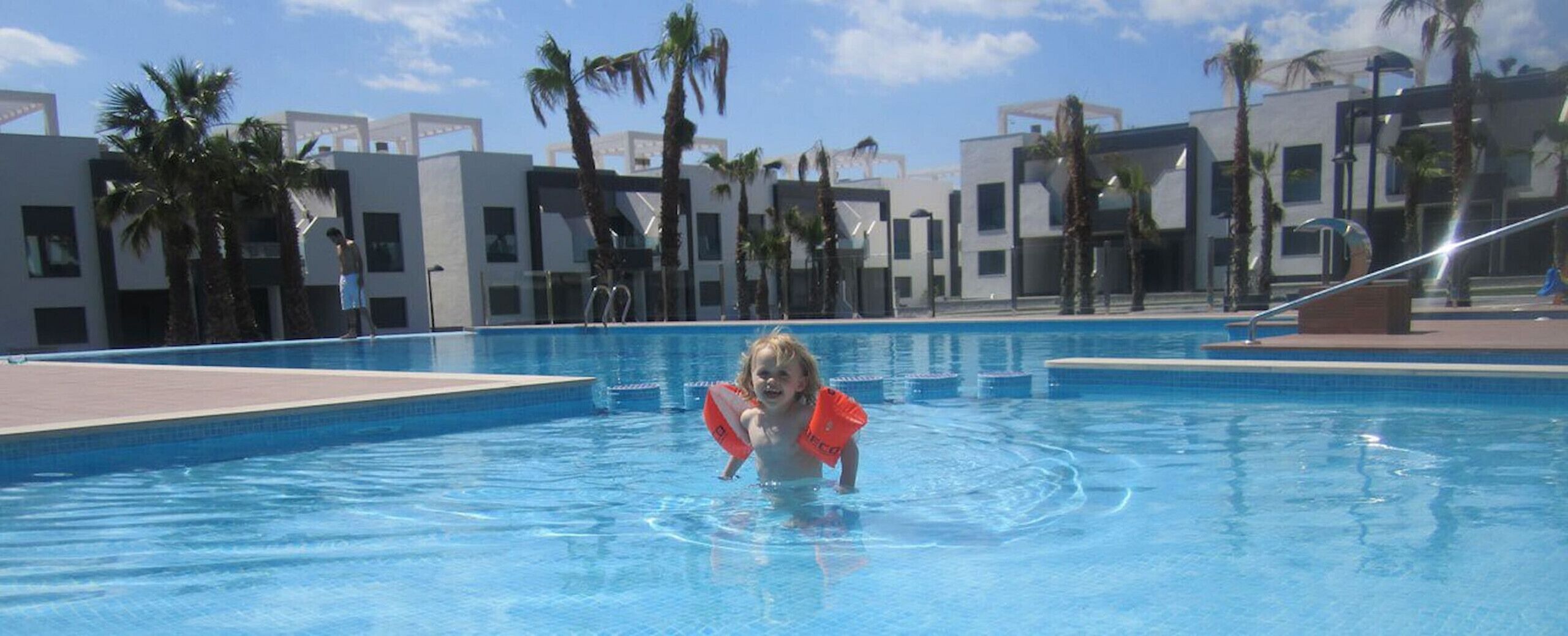 Un niño en una piscina con edificios modernos y palmeras al fondo.