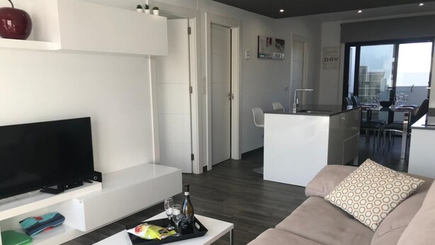 Una sala de estar moderna con una televisión de pantalla plana, un sofá, una mesa de centro con aperitivos y bebidas, y vistas de la ciudad a través de amplias ventanas.