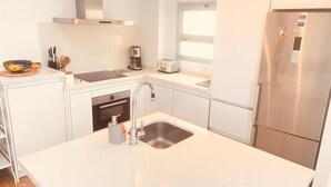 Eine moderne Küche mit weißen Schränken, einem Edelstahlkühlschrank, einem eingebauten Ofen, einer Spüle und einem Fenster über der Arbeitsplatte.
