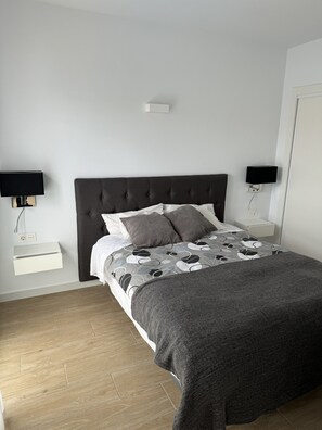Une chambre à coucher avec une tête de lit sombre, un lit recouvert d’une literie à motifs, deux tables de chevet avec des lampes et une commode blanche placée à côté du lit.