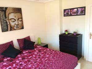 Une chambre à coucher avec un lit, une commode, une petite table sur laquelle est posé un vase, et des œuvres d’art accrochées au mur.