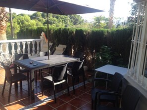 Un patio al aire libre con una mesa y sillas, rodeado por un balcón blanco y abundante vegetación.