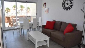 A living room with a brown sofa, white coffee table, and red cushions. There is a dining area with white chairs and a table. A large window provides a view of palm trees outside.