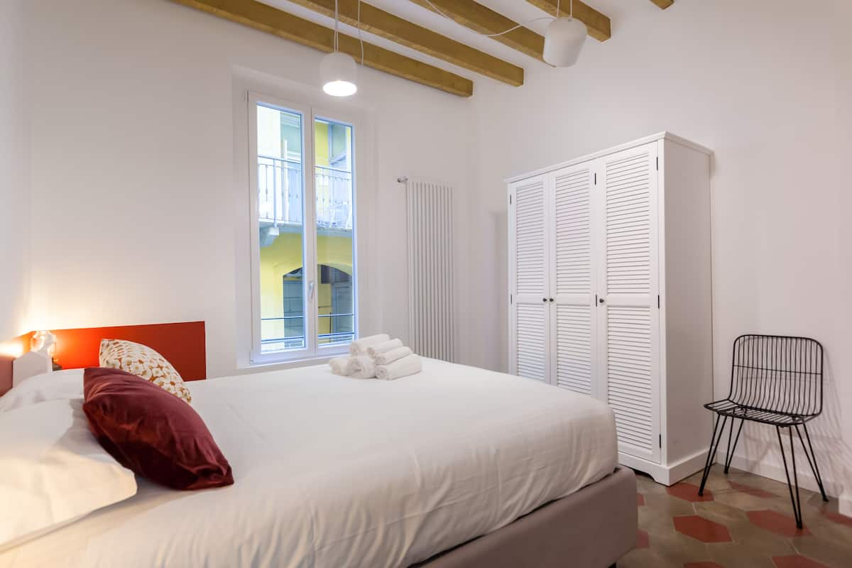 A bedroom with a bed, white curtains, a radiator, a window, and a chair.