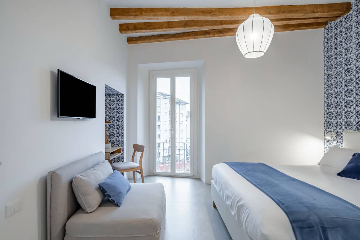 A modern bedroom with a sofa, a bed, a TV, and a balcony with patterned curtains.
