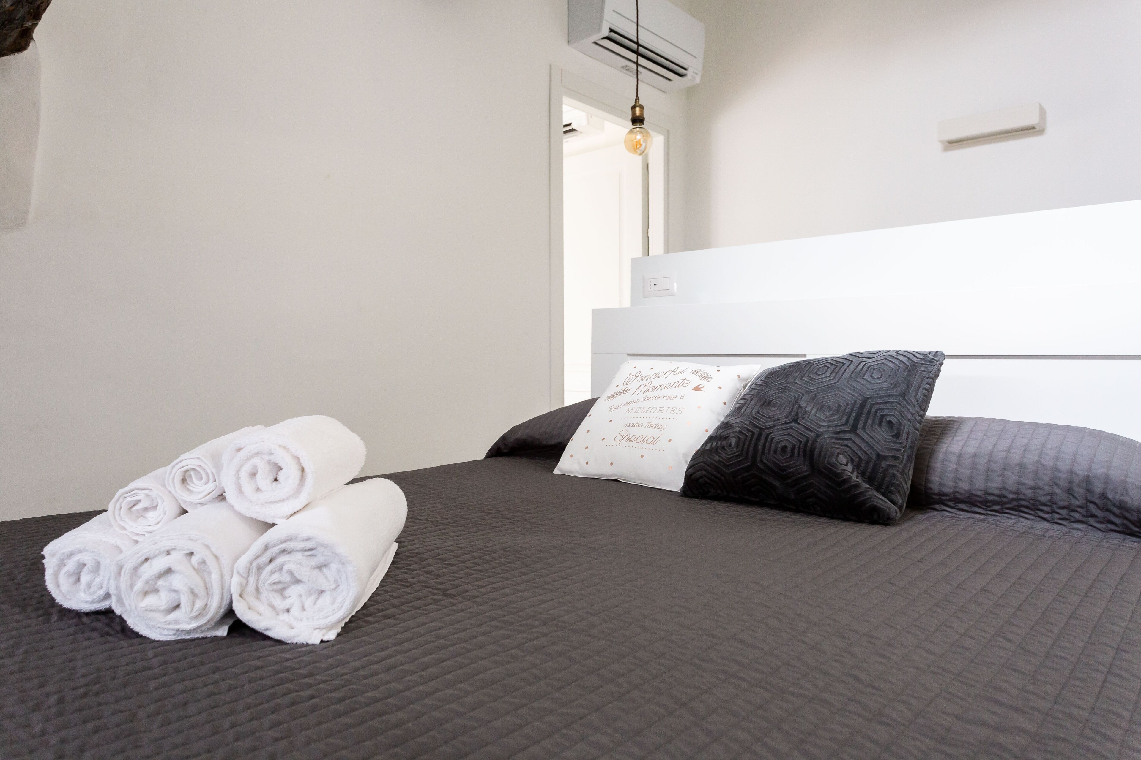 A neatly made bed with rolled white towels, a dark gray quilted bedspread, and a pillow with a geometric pattern.