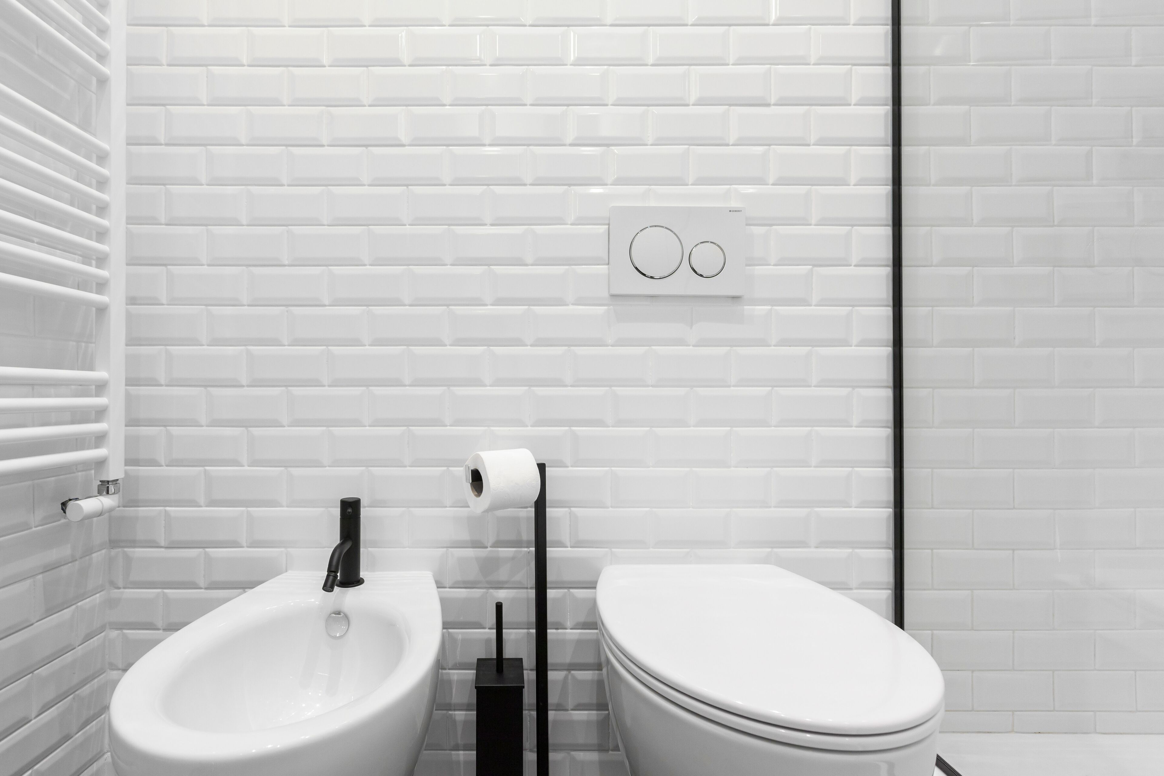 Une salle de bain moderne avec des carreaux de métro blancs, un bidet blanc, des toilettes et un pommeau de bidet noir.