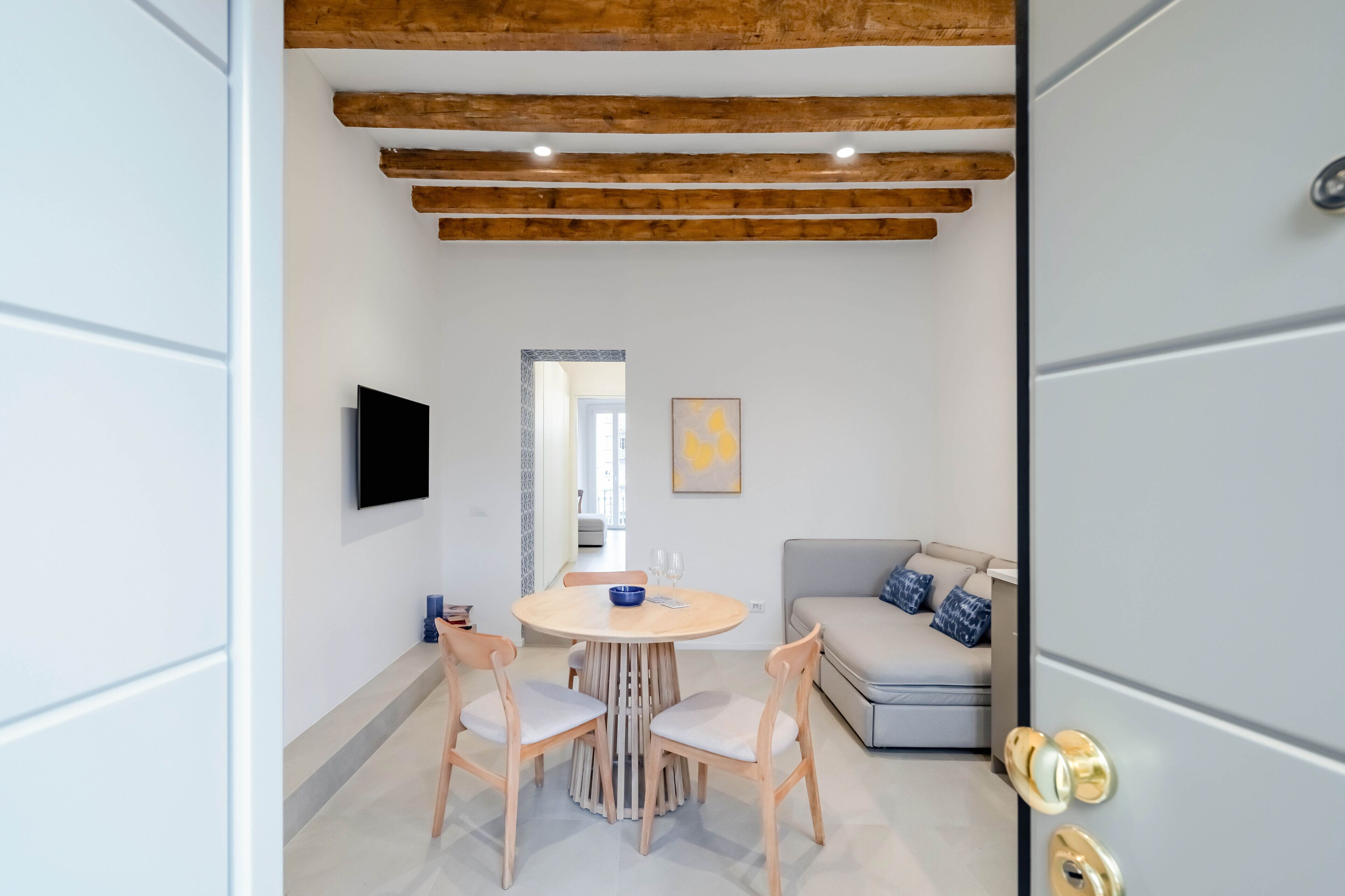 A modern dining area with a round wooden table, four chairs, a TV mounted on the wall, and a sofa with cushions.