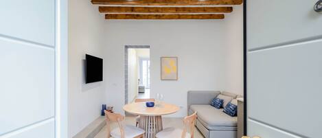 A modern dining area with a round wooden table, four chairs, a TV mounted on the wall, and a sofa with cushions.