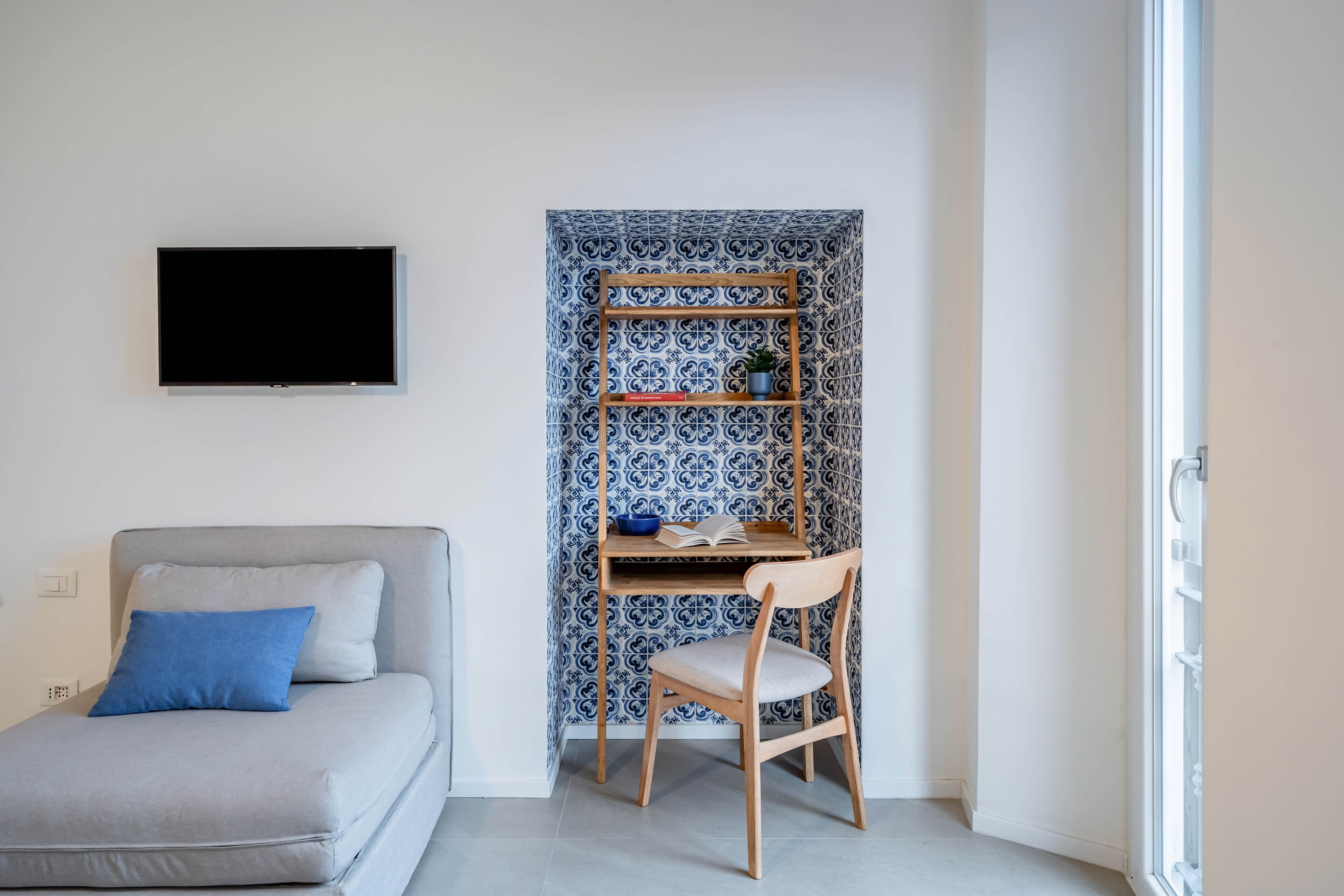 A room with a grey sofa, a wooden chair, a TV, and a bookshelf against a patterned wall.