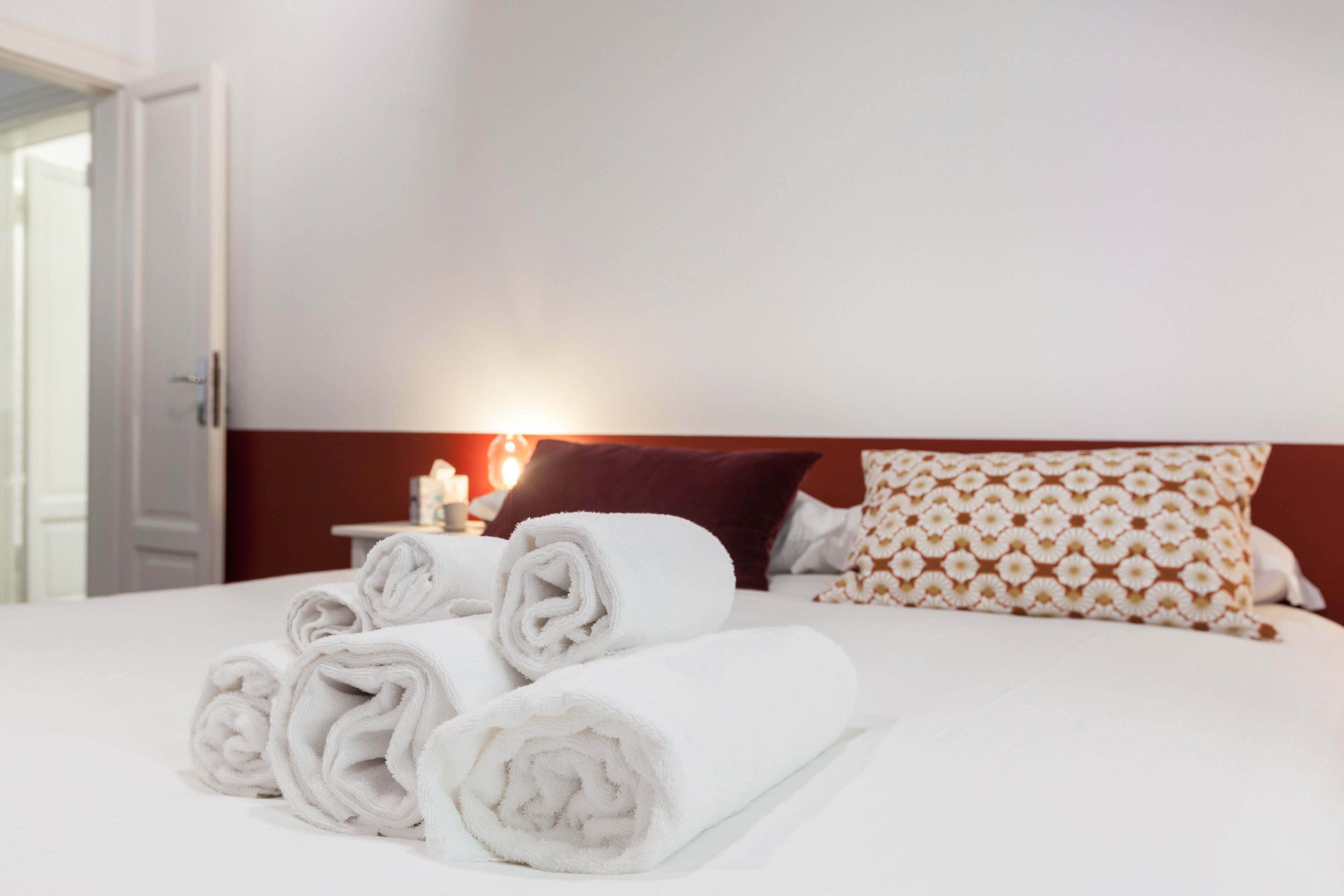 A neatly made bed with rolled white towels, a patterned pillow, and a bedside table with a candle.