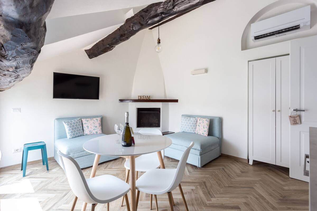 A modern living room with a herringbone floor, a round dining table with chairs, a sofa with patterned cushions, a mounted flat-screen TV, and a built-in fireplace.