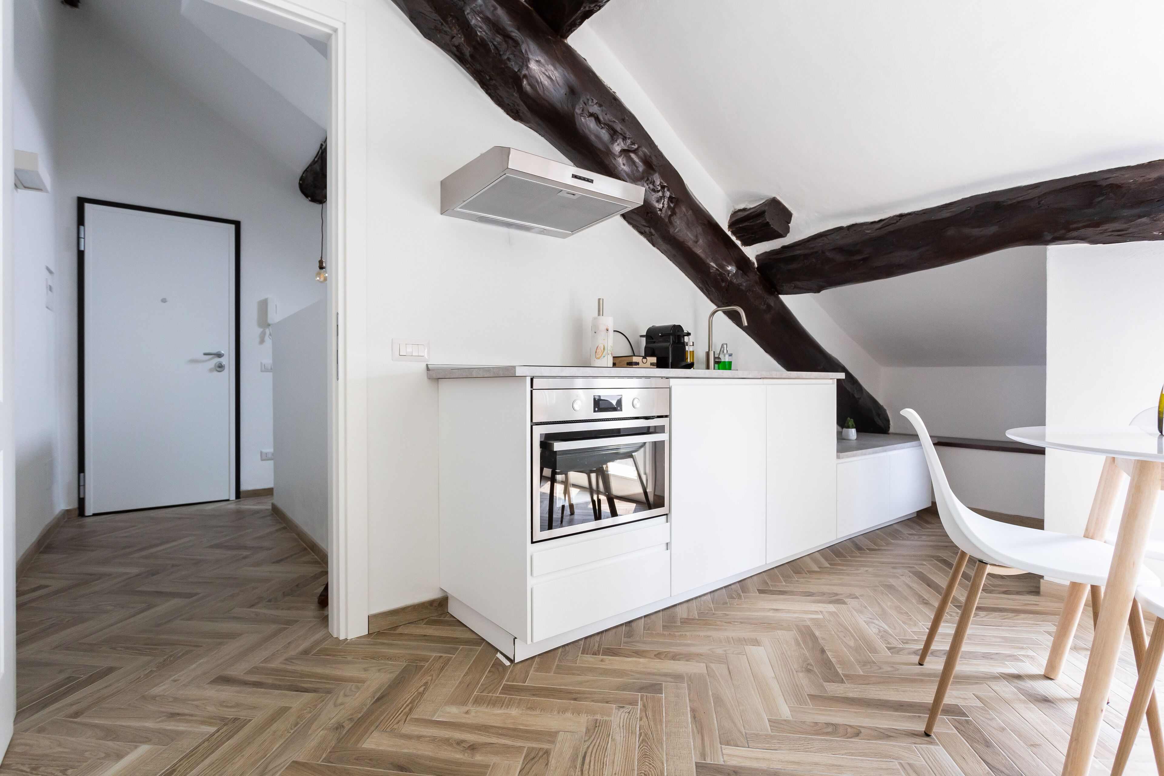 A modern kitchen with white cabinetry, a built-in oven, and a white countertop. There is a wooden ceiling beam and a white door in the background.