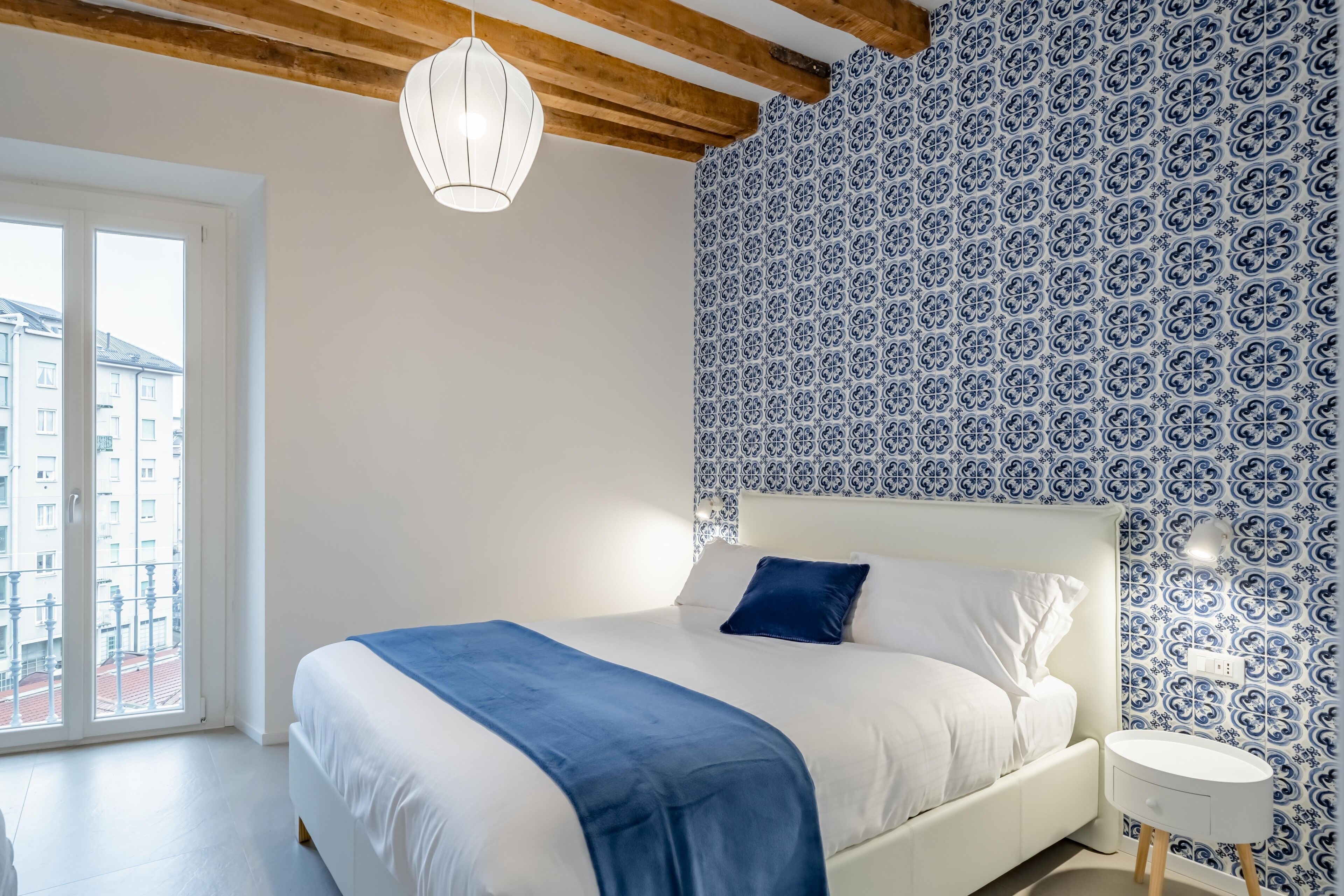 A bedroom with a large bed, a blue throw blanket, a white bedside table, and a patterned wall.