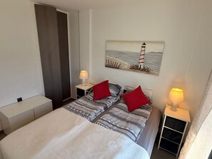 Una habitación de hotel con una cama, dos mesitas de noche, un velador, un cuadro de un faro y una cortina.