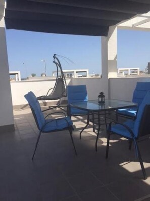 Una terraza en la azotea con una mesa redonda y sillas azules, un sillón colgante y vistas despejadas del horizonte urbano.