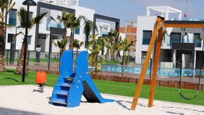 A playground with a blue slide and swings, surrounded by palm trees and modern buildings. - Penthouse Oasis beach IV La zenia (La Zenia)