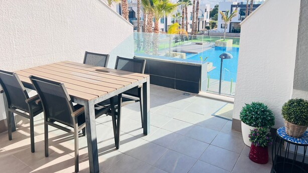 Eine Terrasse mit Holztisch und -stühlen, einer Glasreling, einem Pool und Palmen.