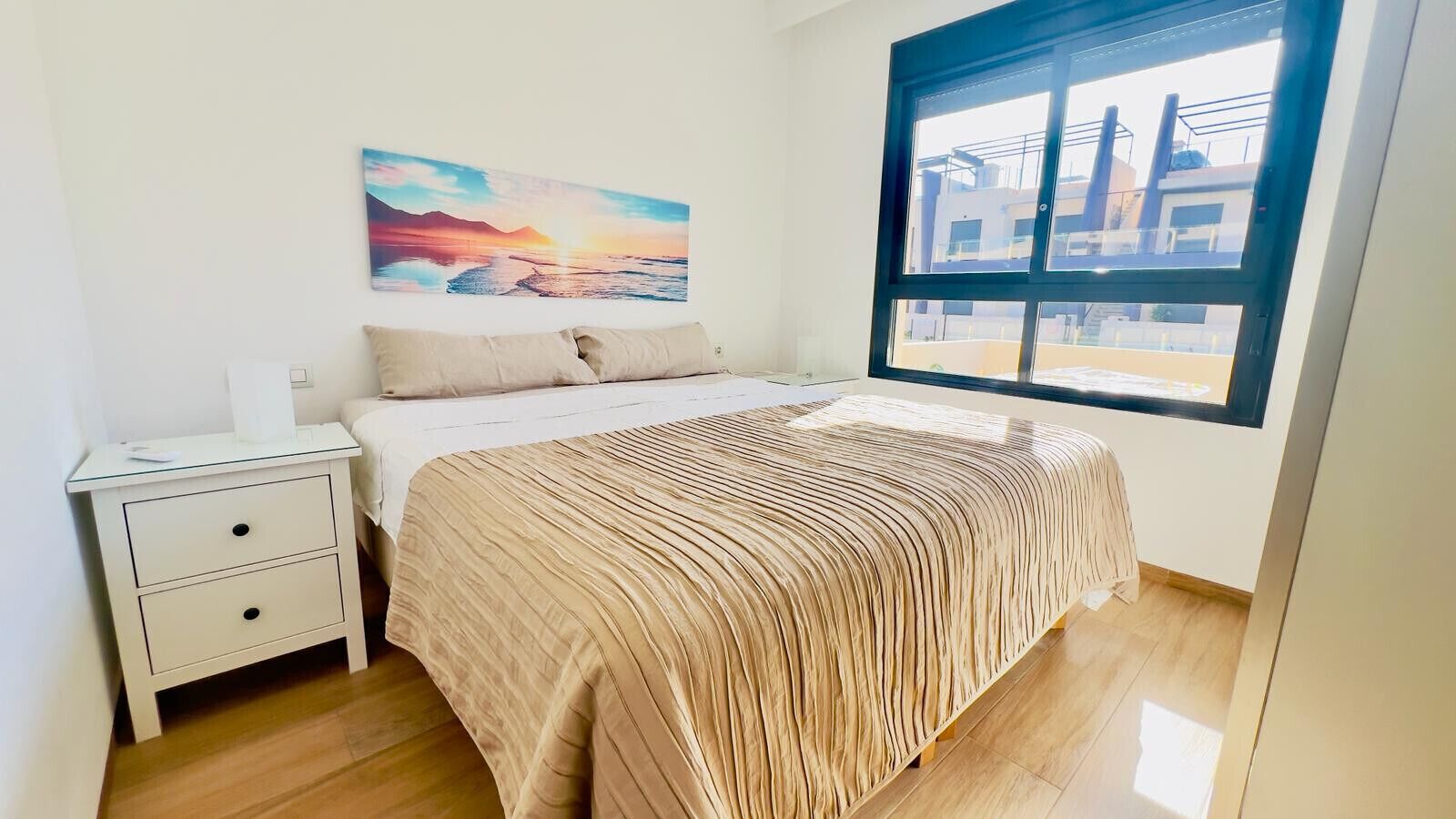 Ein Schlafzimmer mit einem Bett, einem Nachttisch, einem Fenster mit Blick auf Gebäude und einem Bild an der Wand.