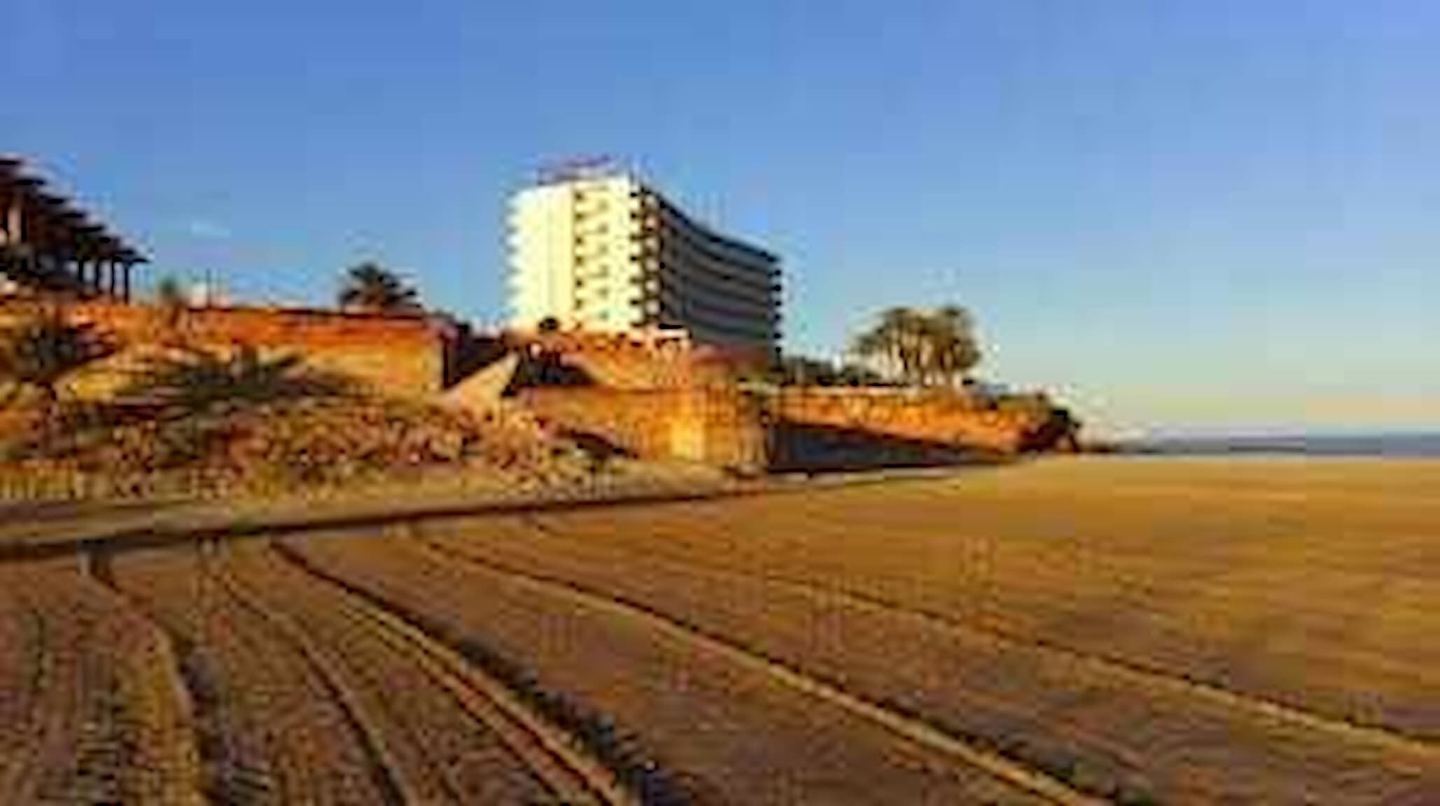 Un hôtel situé sur la côte, avec vue sur la plage, se distinguant par un bâtiment imposant à plusieurs étages et doté d’un toit plat.