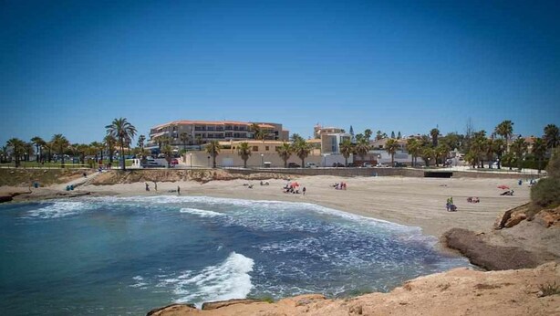 A beach with a large building in the background, palm trees, and a clear sky. - Apartment Playa Flamenca  resd. Playa Terrazas (Orihuela Costa)