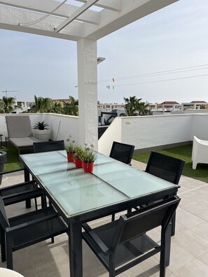 Une terrasse extérieure avec une table à plateau de verre, des chaises noires et des plantes en pot rouges.
