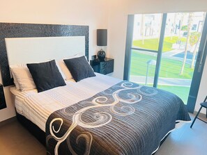 Relax in a spacious room with a large bed, nightstand, and a window offering a view of the outdoor area with lush greenery.