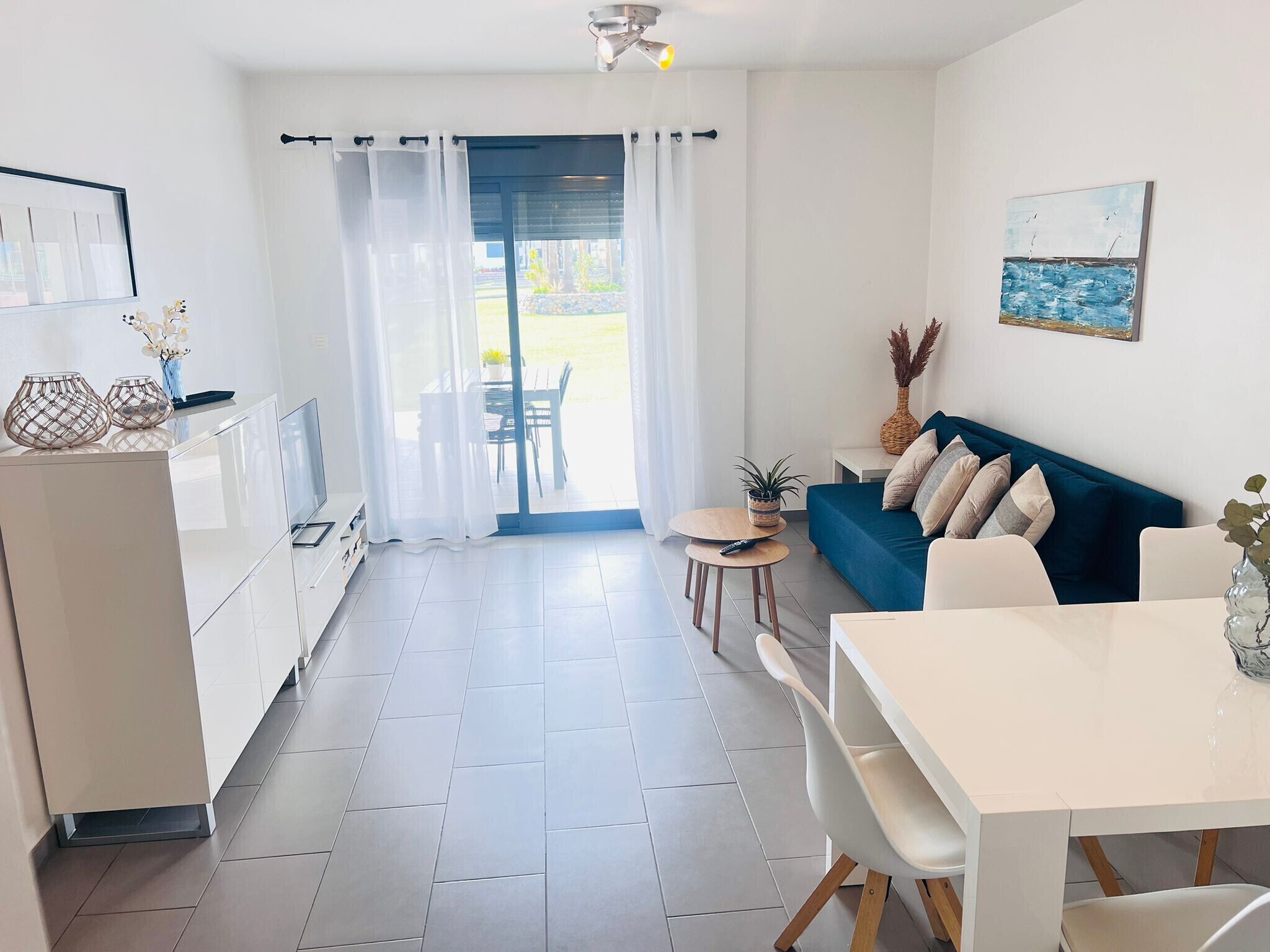 A modern kitchen with white cabinets, a dining area with a white table and chairs, a blue sofa, and a view through a glass door to an outdoor patio.