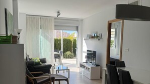 Un salon moderne avec un canapé, une table basse et un téléviseur. On y trouve également un coin repas avec une table et des chaises. La pièce est dotée d’une grande porte-fenêtre coulissante qui donne sur un balcon.